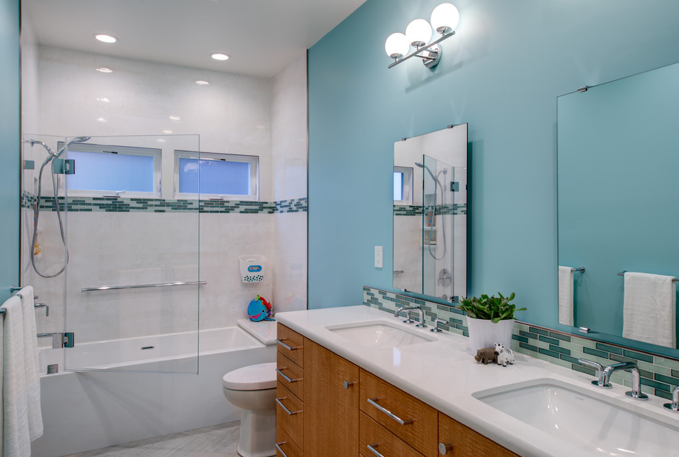 Свежая идея для дизайна: детская ванная комната среднего размера в современном стиле с плоскими фасадами, светлыми деревянными фасадами, ванной в нише, душем над ванной, унитазом-моноблоком, белой плиткой, керамогранитной плиткой, синими стенами, полом из керамогранита, врезной раковиной, столешницей из искусственного кварца, душем с распашными дверями, белой столешницей, нишей, сиденьем для душа, тумбой под две раковины и подвесной тумбой - отличное фото интерьера