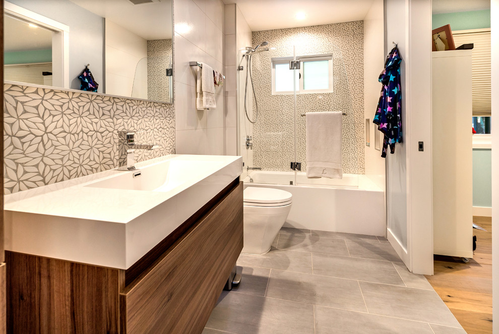 Inredning av ett modernt badrum, med ett integrerad handfat, släta luckor, skåp i mellenmörkt trä, ett badkar i en alkov, en dusch/badkar-kombination, en toalettstol med separat cisternkåpa, blå väggar, grå kakel och vit kakel