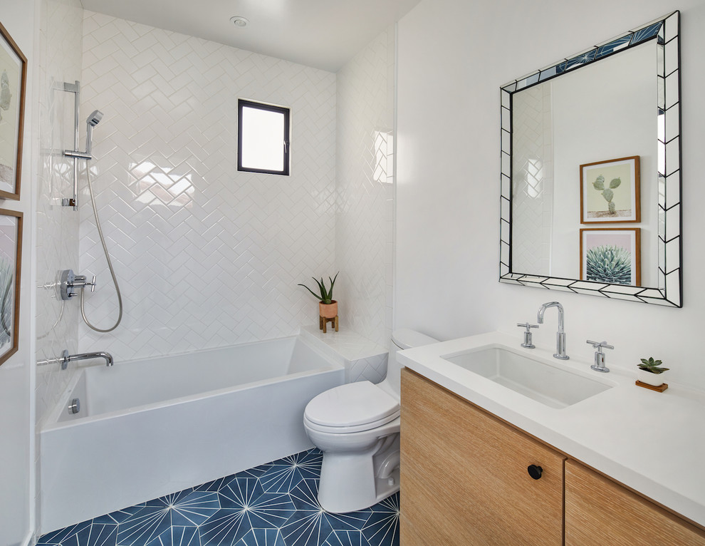 Modern inredning av ett litet vit vitt badrum för barn, med släta luckor, skåp i ljust trä, ett badkar i en alkov, en dusch/badkar-kombination, en toalettstol med hel cisternkåpa, vit kakel, keramikplattor, vita väggar, cementgolv, ett undermonterad handfat, bänkskiva i kvarts och blått golv