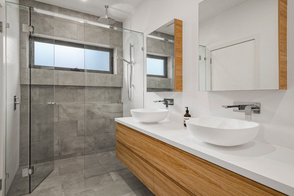 Modern inredning av ett vit vitt badrum, med släta luckor, skåp i mellenmörkt trä, en kantlös dusch, grå kakel, vita väggar, ett fristående handfat, grått golv och dusch med gångjärnsdörr