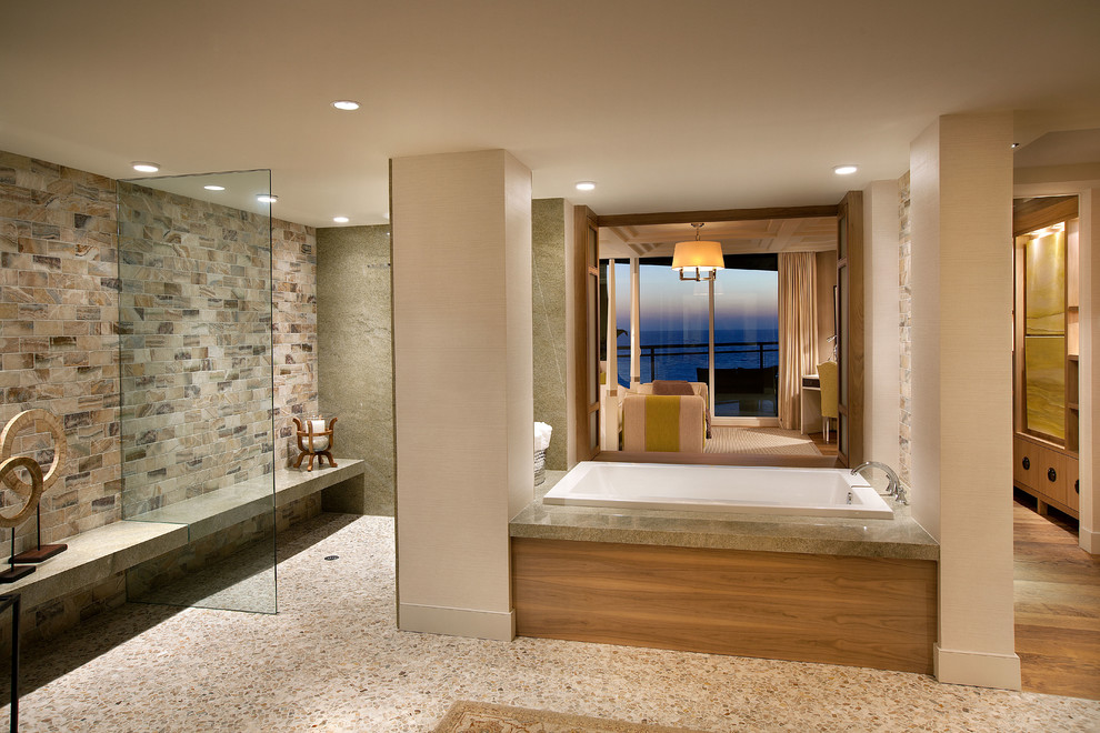 Cette image montre une salle de bain marine avec une baignoire posée, une douche ouverte, un carrelage gris, un carrelage de pierre, un mur gris, un sol en galet, un sol beige et aucune cabine.