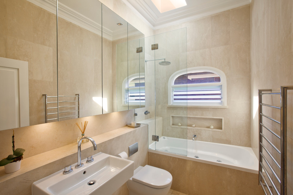 シドニーにある高級なトランジショナルスタイルのおしゃれな浴室 (壁付け型シンク、フラットパネル扉のキャビネット、アルコーブ型浴槽、シャワー付き浴槽	、壁掛け式トイレ、ベージュのタイル、石タイル、ベージュの壁) の写真