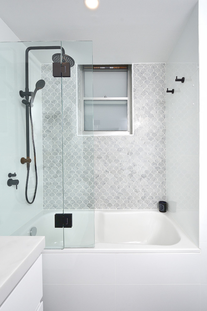 Modern inredning av ett litet vit vitt en-suite badrum, med luckor med glaspanel, vita skåp, ett badkar i en alkov, en öppen dusch, en toalettstol med hel cisternkåpa, vit kakel, keramikplattor, vita väggar, cementgolv, ett fristående handfat, bänkskiva i akrylsten, svart golv och med dusch som är öppen