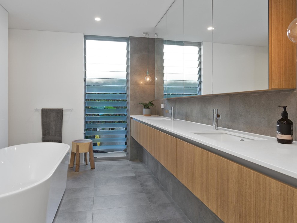 Modern ensuite bathroom in Perth with flat-panel cabinets, medium wood cabinets, a freestanding bath, grey tiles, white walls, a submerged sink, grey floors and white worktops.