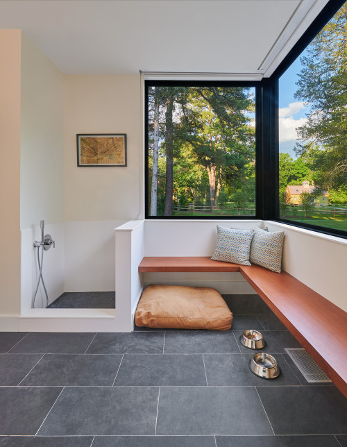 Bright mudroom with an orange bench and large black framed windows looking out on trees.  There are gray tile floors with an orange dog bed and a white tiled dog shower.  