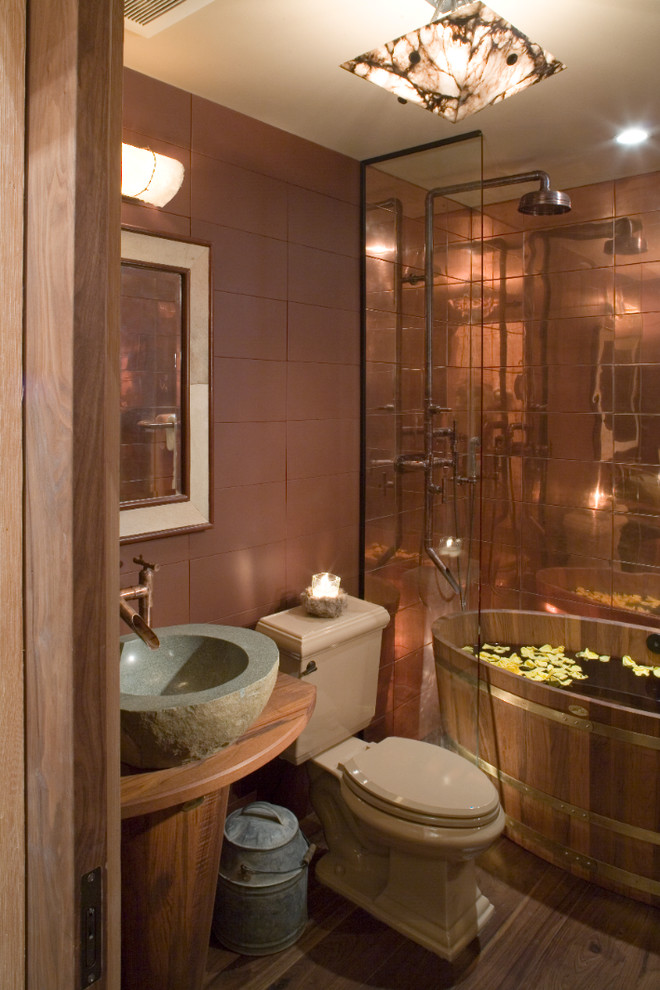 Rustik inredning av ett badrum, med brun kakel, mörkt trägolv, ett fristående handfat, ett fristående badkar och en toalettstol med separat cisternkåpa