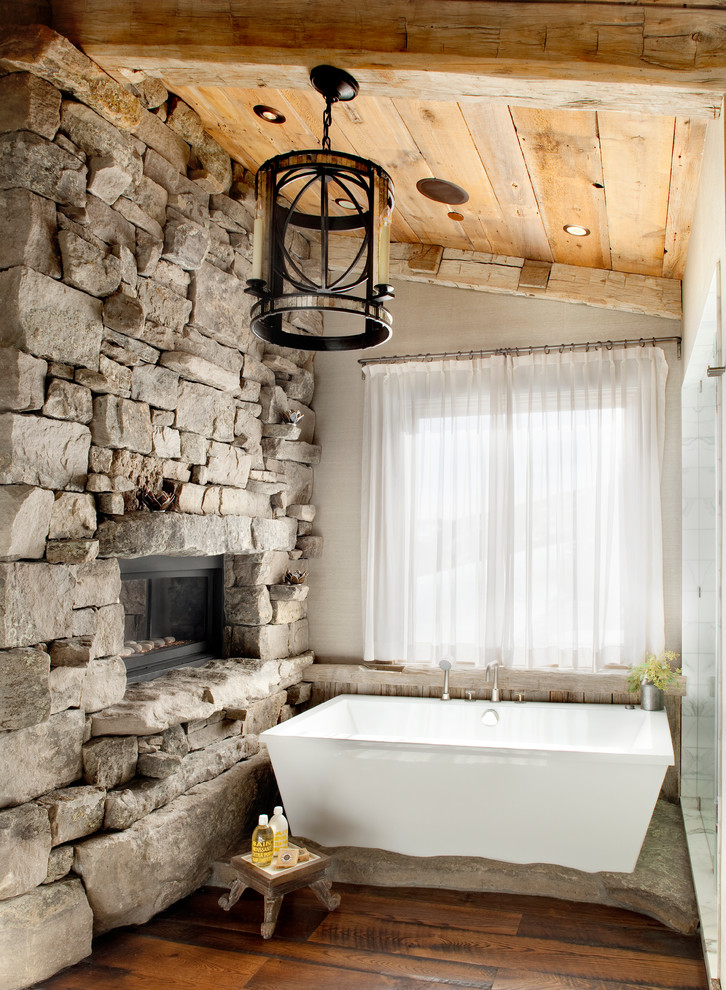 Modelo de cuarto de baño rústico con bañera exenta, paredes grises, suelo de madera oscura y piedra