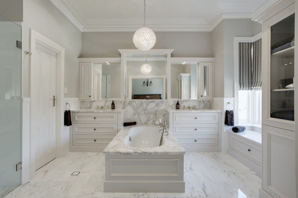 Идея дизайна: большая главная ванная комната в классическом стиле с мраморной столешницей, полновстраиваемой ванной, врезной раковиной, фасадами с утопленной филенкой, белыми фасадами, серыми стенами, мраморным полом, серой плиткой, плиткой из листового камня и серой столешницей