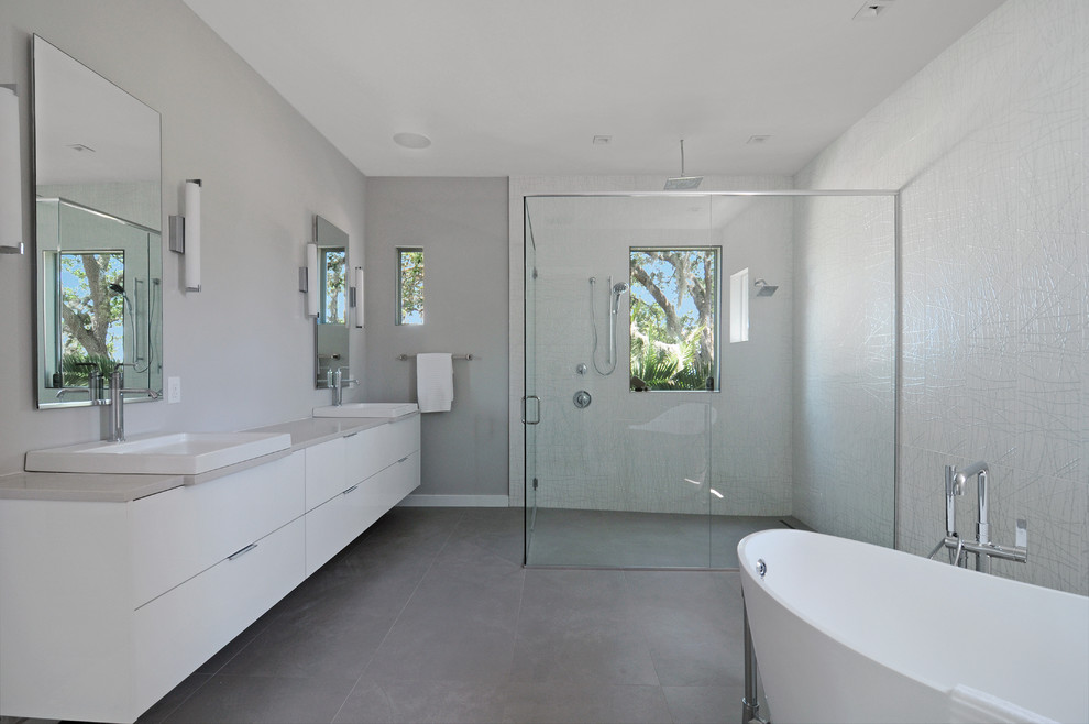Modern inredning av ett badrum, med släta luckor, vita skåp, ett fristående badkar, en kantlös dusch, grå kakel, grå väggar, ett avlångt handfat och dusch med gångjärnsdörr