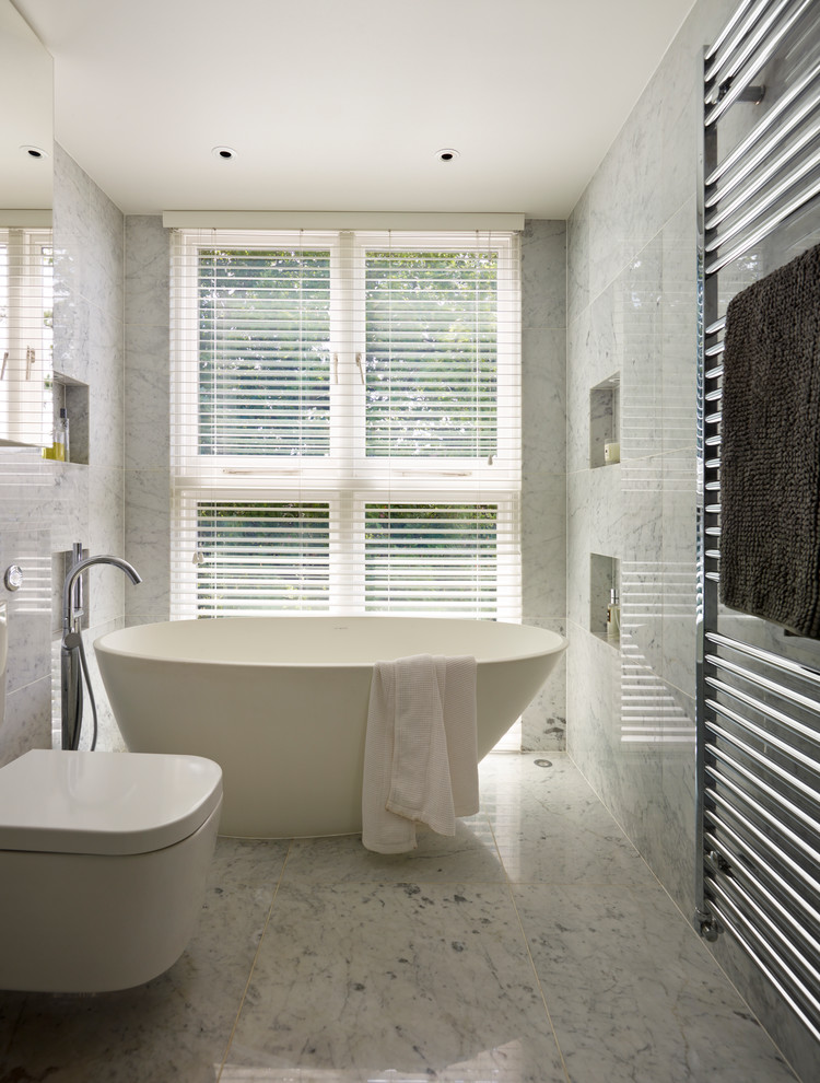 Inspiration for a contemporary bathroom in London with a freestanding bath, a wall mounted toilet, grey tiles, grey walls, marble flooring and marble tiles.