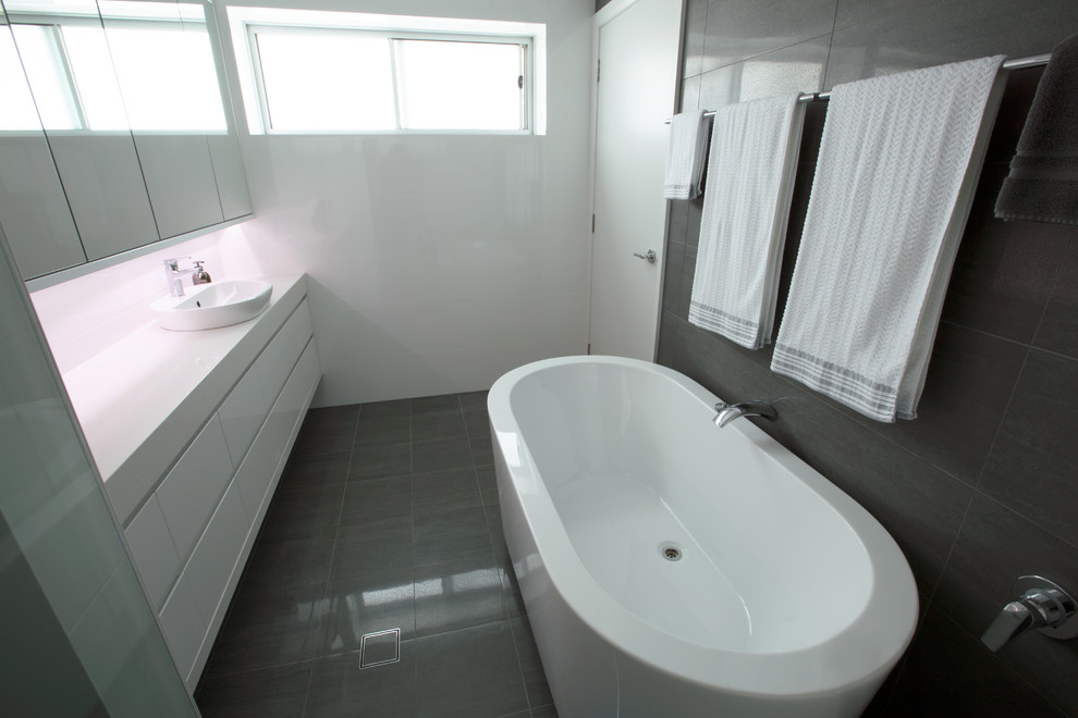 Exemple d'une salle de bain principale tendance avec une vasque, un placard à porte plane, des portes de placard blanches, un plan de toilette en quartz modifié, une baignoire indépendante, un carrelage gris, des carreaux de porcelaine, un mur blanc et un sol en carrelage de porcelaine.