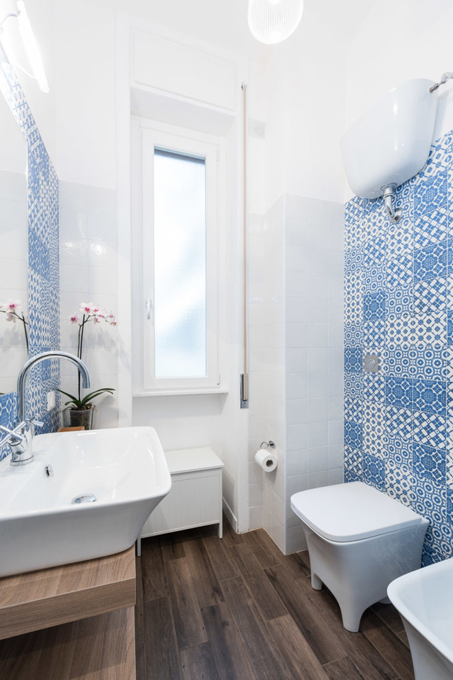 Bild på ett litet maritimt badrum, med ett konsol handfat, öppna hyllor, skåp i mellenmörkt trä, blå kakel, keramikplattor och en toalettstol med hel cisternkåpa