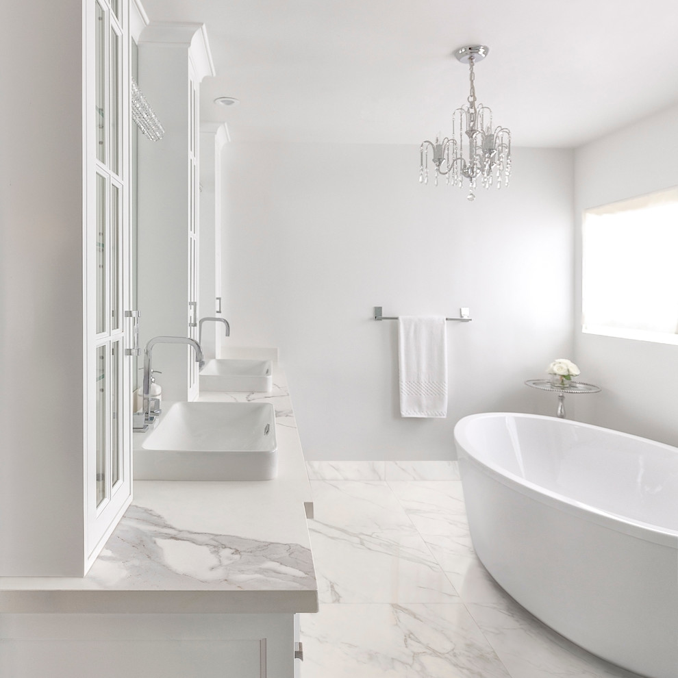 エセックスにあるコンテンポラリースタイルのおしゃれなマスターバスルーム (ガラス扉のキャビネット、白いキャビネット、置き型浴槽、白い壁、大理石の床、オーバーカウンターシンク、大理石の洗面台、白い床) の写真