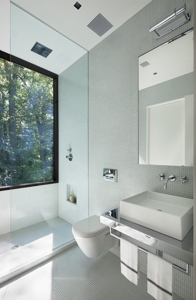Foto på ett stort funkis badrum, med ett väggmonterat handfat, bänkskiva i rostfritt stål, en vägghängd toalettstol, vit kakel, glaskakel, vita väggar och mörkt trägolv