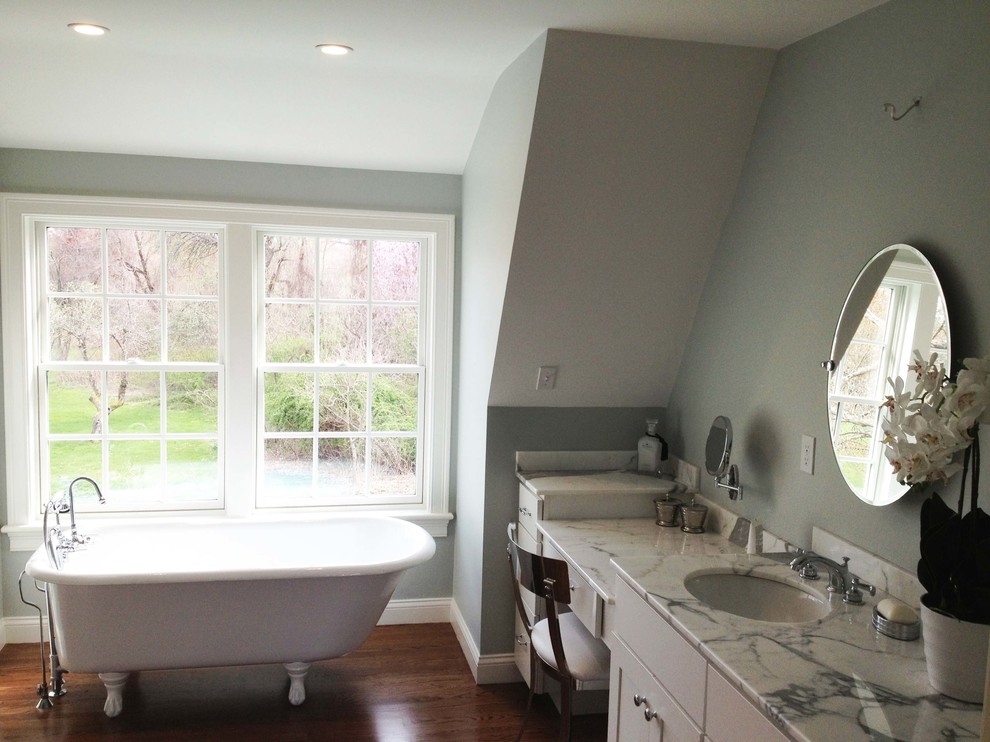 Exemple d'une grande salle de bain principale chic avec un placard à porte shaker, des portes de placard blanches, une baignoire sur pieds, un mur gris, un sol en bois brun, un lavabo encastré, un plan de toilette en marbre et un sol marron.