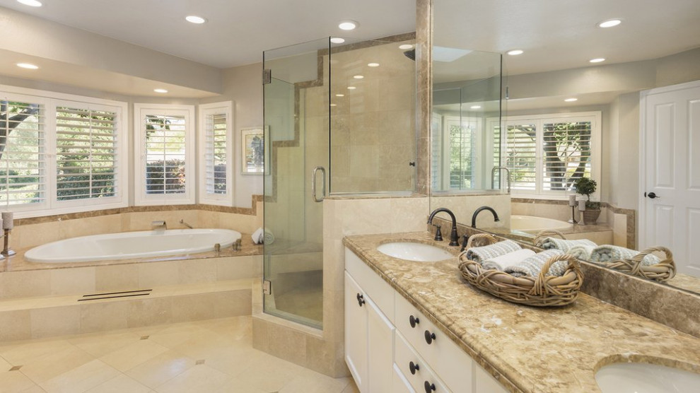 На фото: главная ванная комната среднего размера в классическом стиле с фасадами с утопленной филенкой, белыми фасадами, полновстраиваемой ванной, угловым душем, раздельным унитазом, коричневой плиткой, плиткой из известняка, бежевыми стенами, полом из известняка, врезной раковиной, столешницей из известняка, бежевым полом, душем с распашными дверями, коричневой столешницей и тумбой под две раковины с