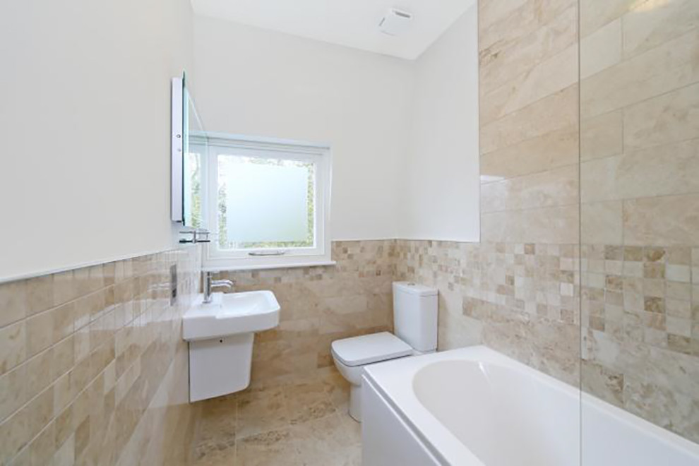Photo of a small contemporary ensuite bathroom in London with a pedestal sink, flat-panel cabinets, white cabinets, an alcove bath, a one-piece toilet, beige tiles, stone tiles, white walls and marble flooring.