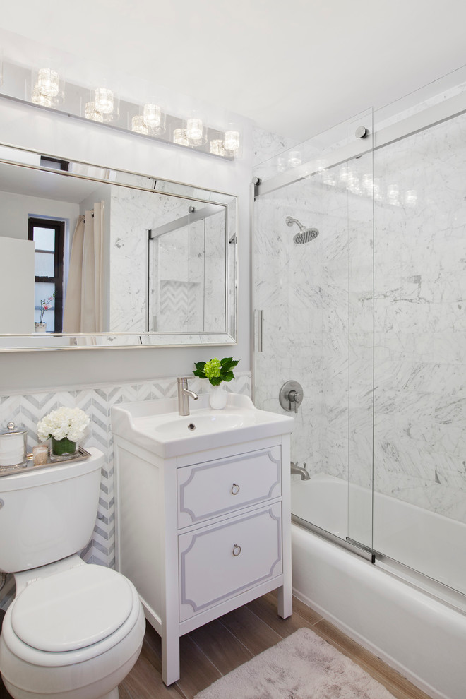 Klassisches Badezimmer mit weißen Schränken, Badewanne in Nische, Duschbadewanne, Wandtoilette mit Spülkasten, weißer Wandfarbe, braunem Holzboden, Waschtischkonsole, weißen Fliesen und flächenbündigen Schrankfronten in New York