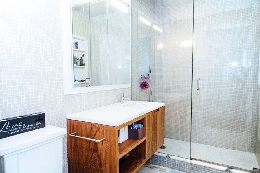 Medium sized traditional ensuite bathroom in New York with flat-panel cabinets, medium wood cabinets, an alcove shower, a two-piece toilet, grey tiles, mosaic tiles, grey walls, mosaic tile flooring, a submerged sink, marble worktops, grey floors, a hinged door and white worktops.