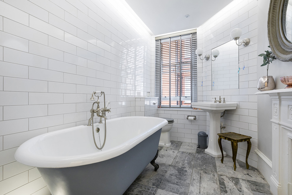 Foto på ett en-suite badrum, med ett badkar med tassar, en toalettstol med separat cisternkåpa, vit kakel, tunnelbanekakel, ett piedestal handfat och grått golv