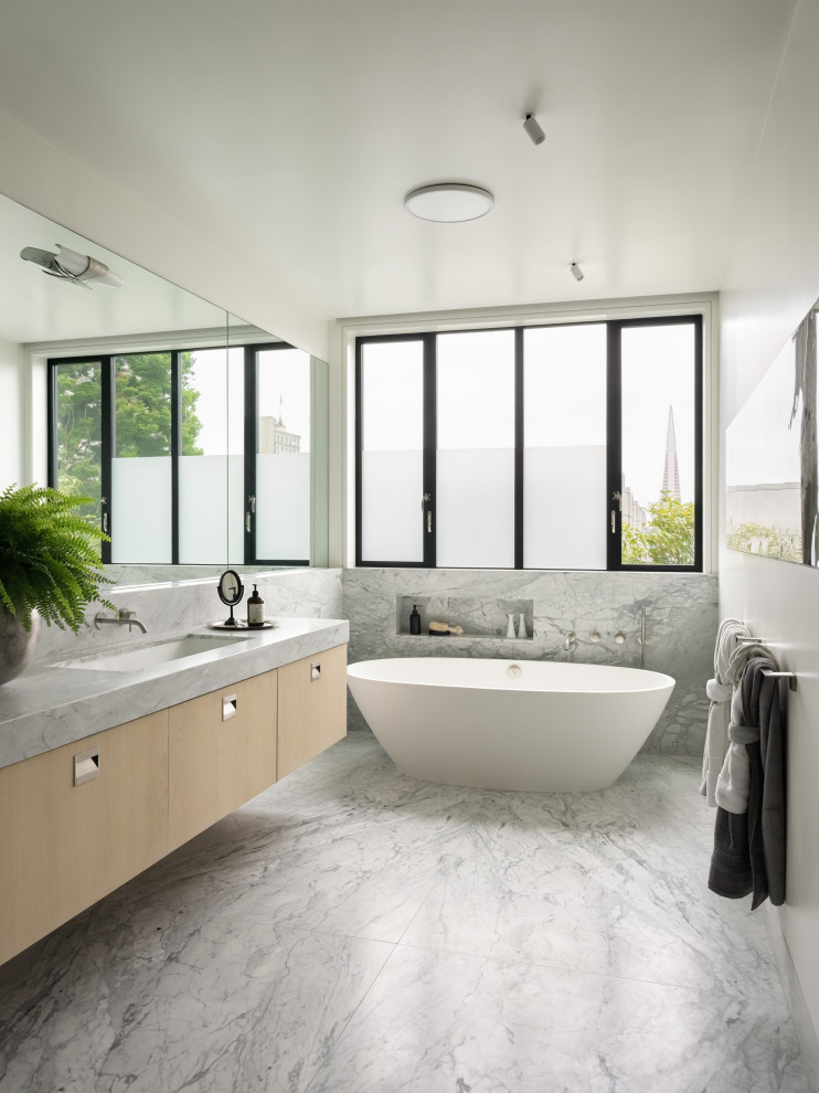 Aménagement d'une salle de bain contemporaine en bois clair avec un placard à porte plane, une baignoire indépendante, un carrelage gris, un mur blanc, un lavabo encastré, un sol gris et un plan de toilette gris.