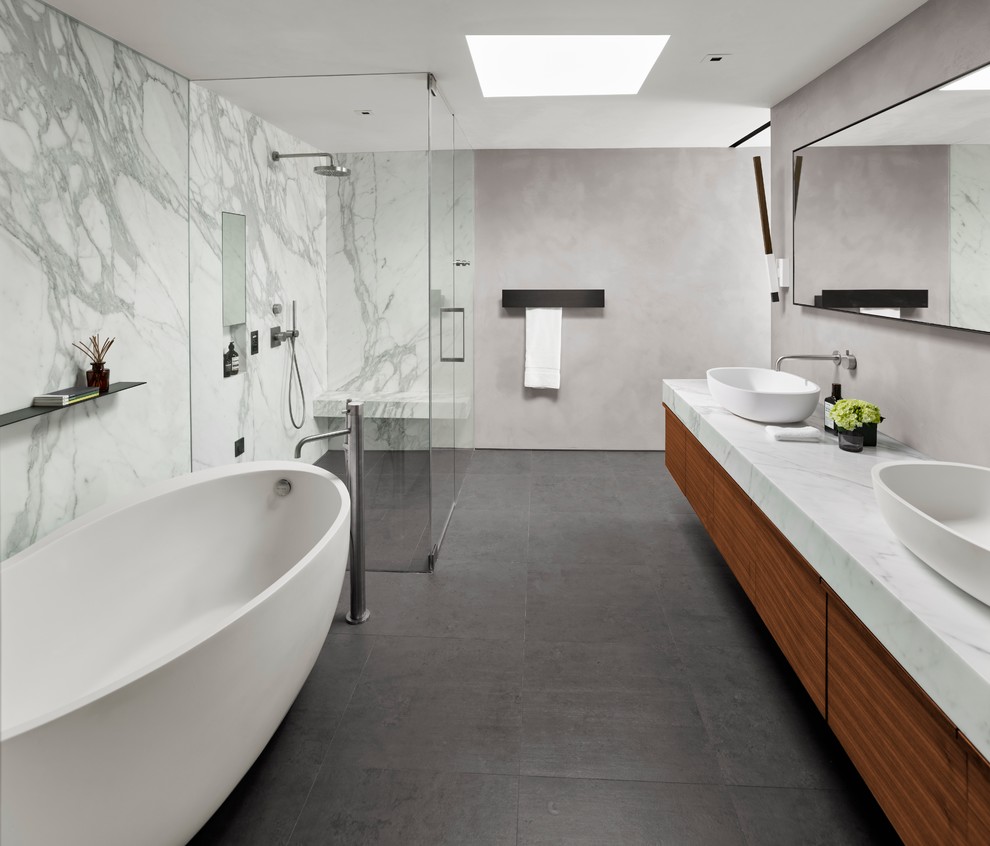 Cette image montre une salle de bain design en bois foncé avec un placard à porte plane, une baignoire indépendante, une douche d'angle, un carrelage blanc, des dalles de pierre, un mur gris et une cabine de douche à porte battante.