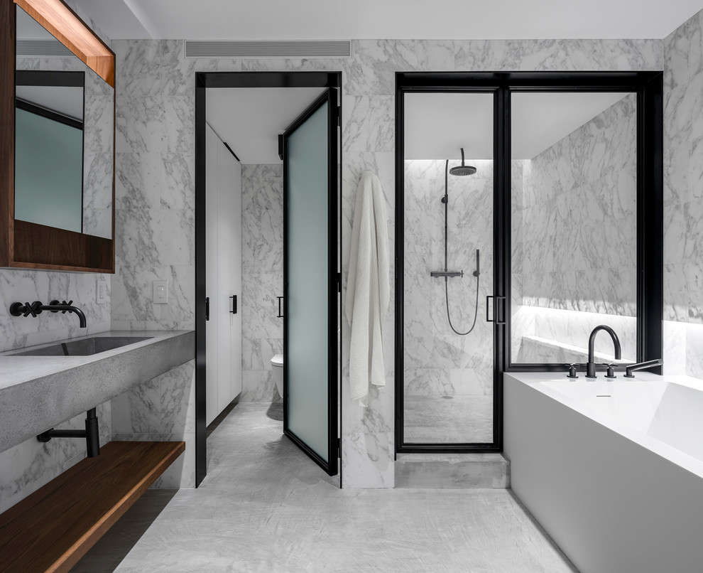 This is an example of a large contemporary ensuite bathroom in New York with marble tiles, concrete flooring, an integrated sink, concrete worktops, grey floors, a hinged door, grey worktops, open cabinets, an alcove shower, grey tiles, grey walls and a freestanding bath.