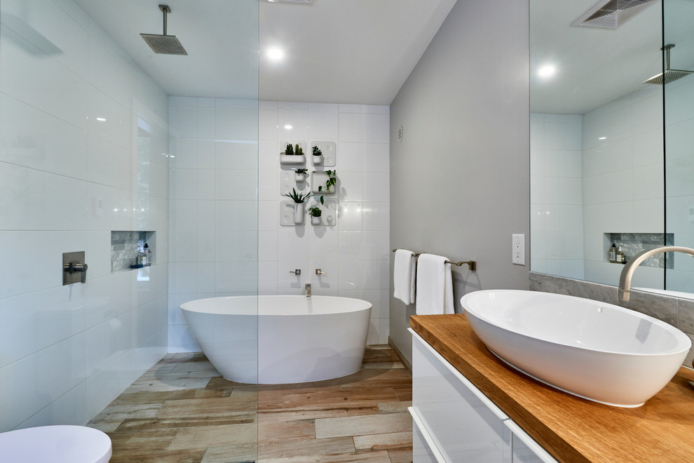 Foto på ett mellanstort skandinaviskt brun en-suite badrum, med släta luckor, vita skåp, ett fristående badkar, en öppen dusch, vit kakel, porslinskakel, grå väggar, klinkergolv i porslin, ett fristående handfat och träbänkskiva