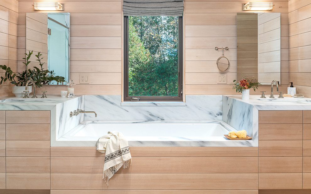 Modelo de cuarto de baño principal rural con puertas de armario de madera clara, bañera encastrada, paredes beige, lavabo bajoencimera, encimera de mármol y armarios con paneles lisos