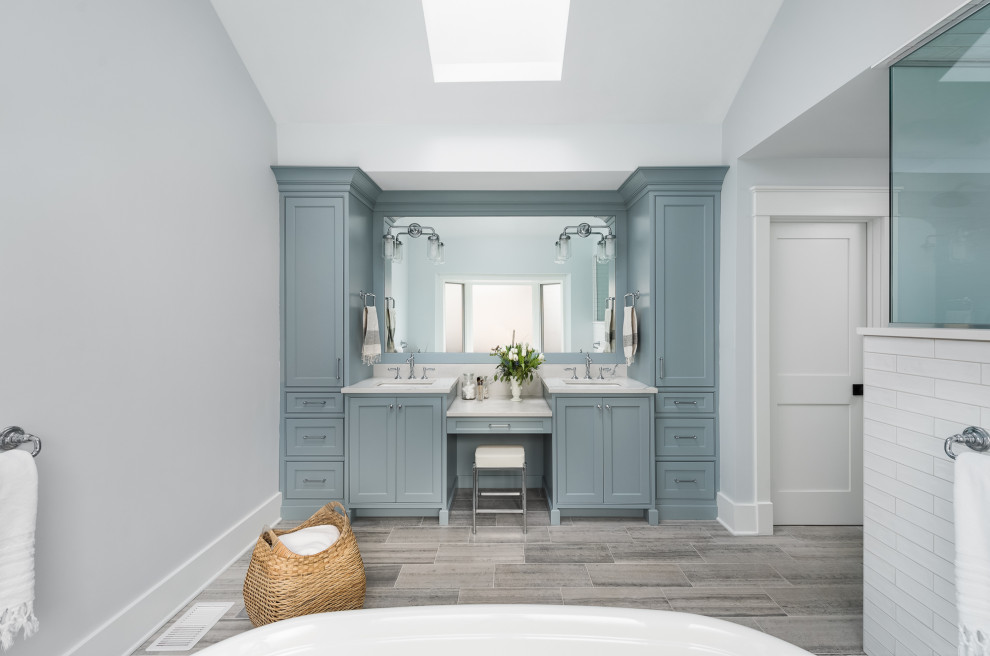 Идея дизайна: большая главная ванная комната в стиле кантри с синими фасадами, отдельно стоящей ванной, угловым душем, раздельным унитазом, белой плиткой, керамогранитной плиткой, белыми стенами, полом из керамогранита, врезной раковиной, столешницей из искусственного кварца, серым полом, душем с распашными дверями, белой столешницей, сиденьем для душа, тумбой под две раковины, встроенной тумбой и фасадами с декоративным кантом