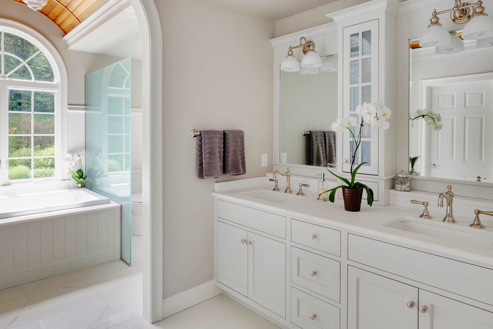 Exemple d'une grande salle de bain principale chic avec des portes de placard blanches, un plan de toilette en marbre, une baignoire posée, des carreaux de céramique, un mur beige, un sol en marbre, un lavabo encastré et un placard à porte plane.