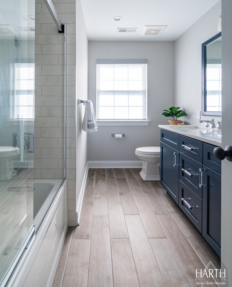 This is an example of a medium sized classic shower room bathroom in Philadelphia with shaker cabinets, blue cabinets, an alcove bath, a shower/bath combination, a two-piece toilet, white tiles, metro tiles, grey walls, porcelain flooring, a submerged sink, engineered stone worktops, beige floors, a sliding door and white worktops.