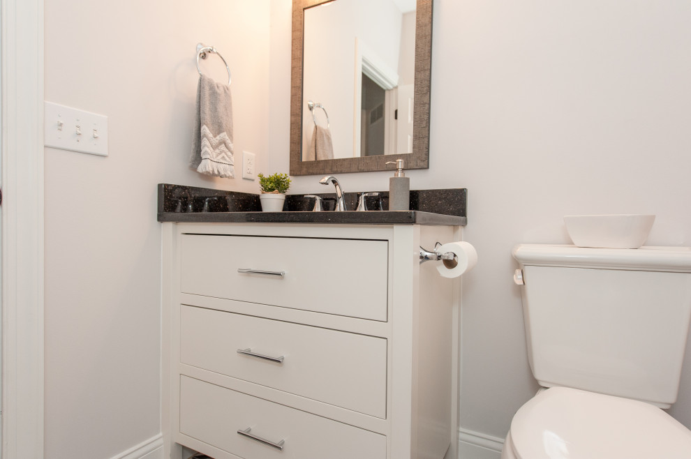 Modelo de cuarto de baño único actual con sanitario de una pieza, paredes blancas, suelo con mosaicos de baldosas, lavabo bajoencimera, encimera de granito, suelo gris y encimeras negras