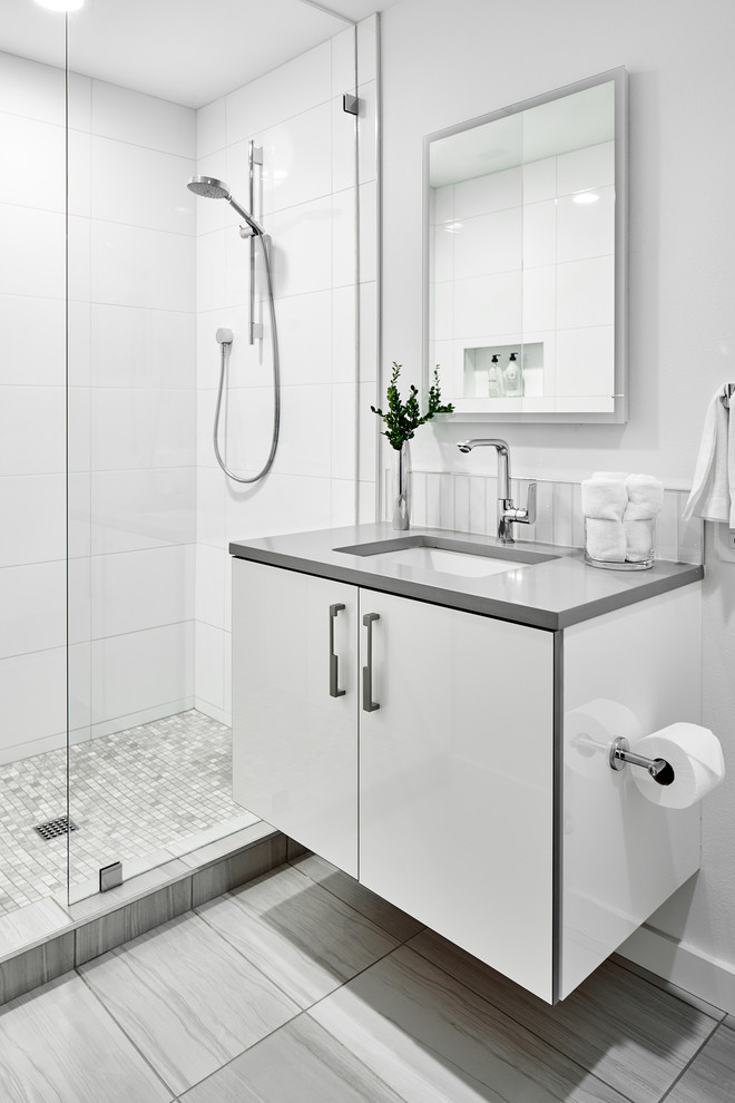 Inredning av ett 60 tals stort grå grått badrum med dusch, med släta luckor, vita skåp, ett badkar i en alkov, en dusch/badkar-kombination, en toalettstol med hel cisternkåpa, vit kakel, porslinskakel, vita väggar, klinkergolv i porslin, ett undermonterad handfat, bänkskiva i kvarts, grått golv och med dusch som är öppen
