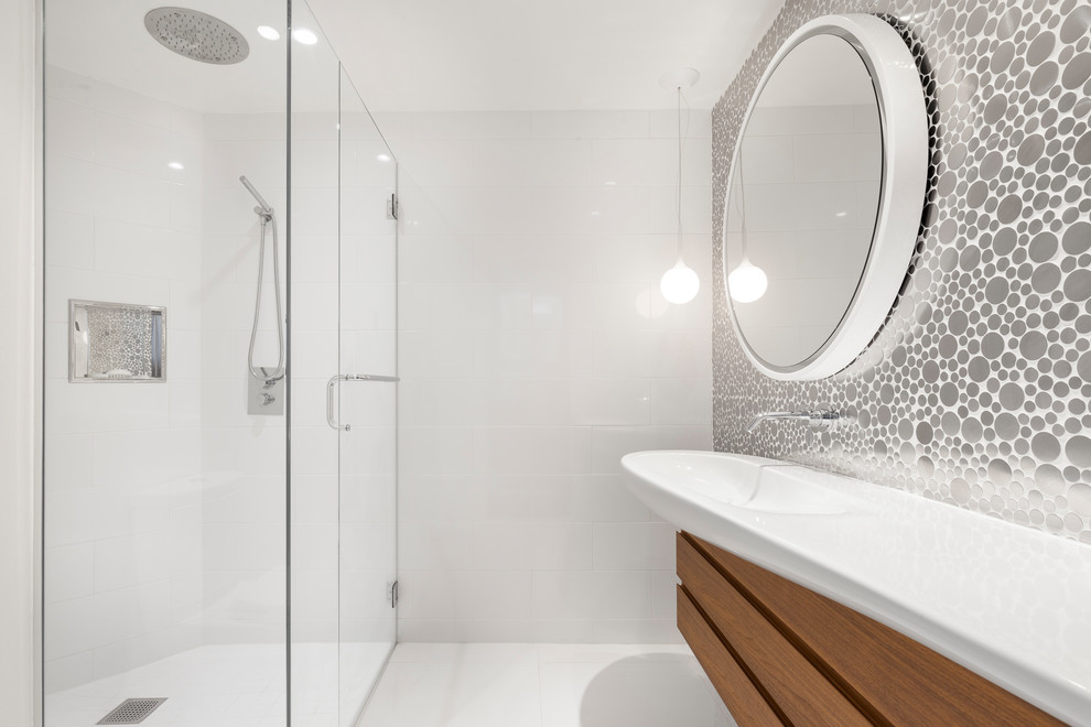 This is an example of a small contemporary family bathroom with flat-panel cabinets, medium wood cabinets, white tiles, solid surface worktops, a built-in shower, a one-piece toilet, white walls, ceramic flooring, a console sink, white floors and a hinged door.