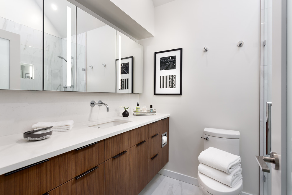 Cette photo montre une salle de bain tendance en bois brun de taille moyenne avec un placard à porte plane, un carrelage blanc, une cabine de douche à porte battante, WC à poser, du carrelage en marbre, un mur blanc, un sol en carrelage de porcelaine, un lavabo encastré, un plan de toilette en quartz, un sol gris et un plan de toilette blanc.