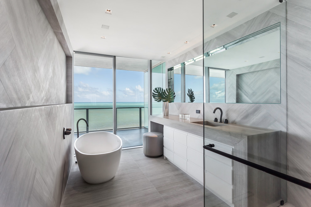 Cette photo montre une salle de bain tendance avec un placard à porte plane, des portes de placard blanches, une baignoire indépendante, un carrelage gris, un lavabo intégré, un sol gris et un plan de toilette gris.