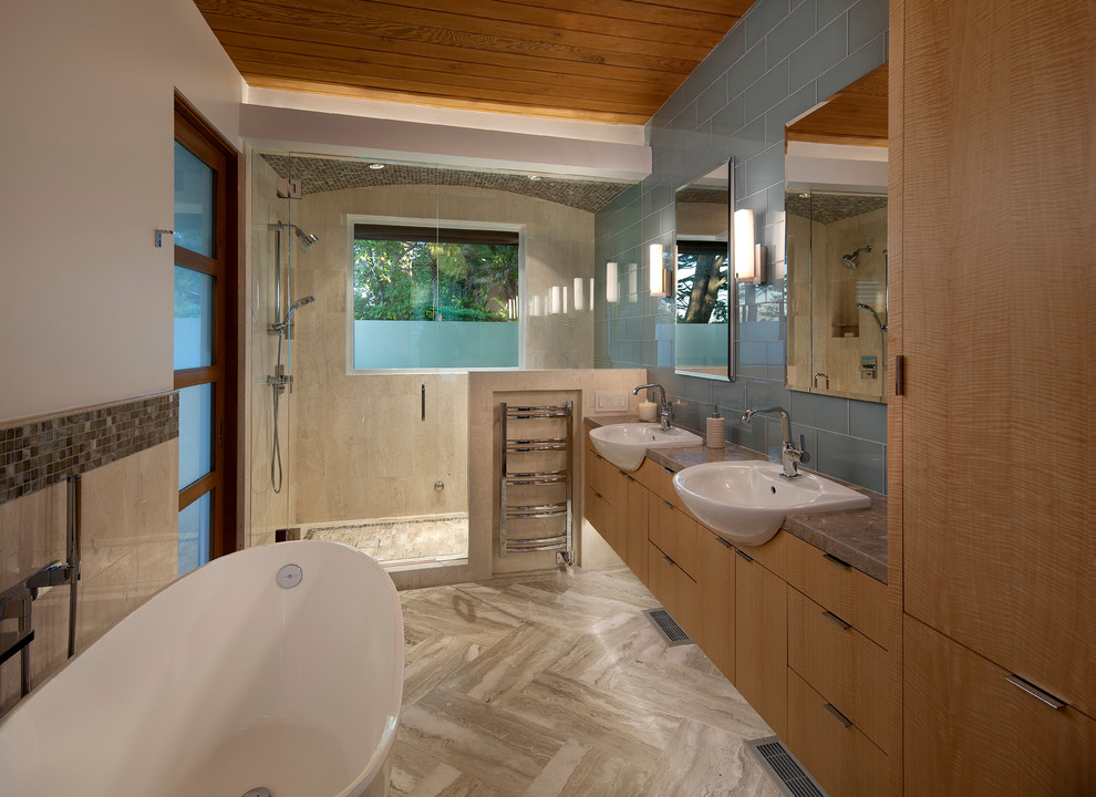 На фото: главная ванная комната среднего размера в стиле модернизм с накладной раковиной, плоскими фасадами, светлыми деревянными фасадами, столешницей из гранита, отдельно стоящей ванной, душем в нише, синей плиткой, стеклянной плиткой, бежевыми стенами и полом из известняка с