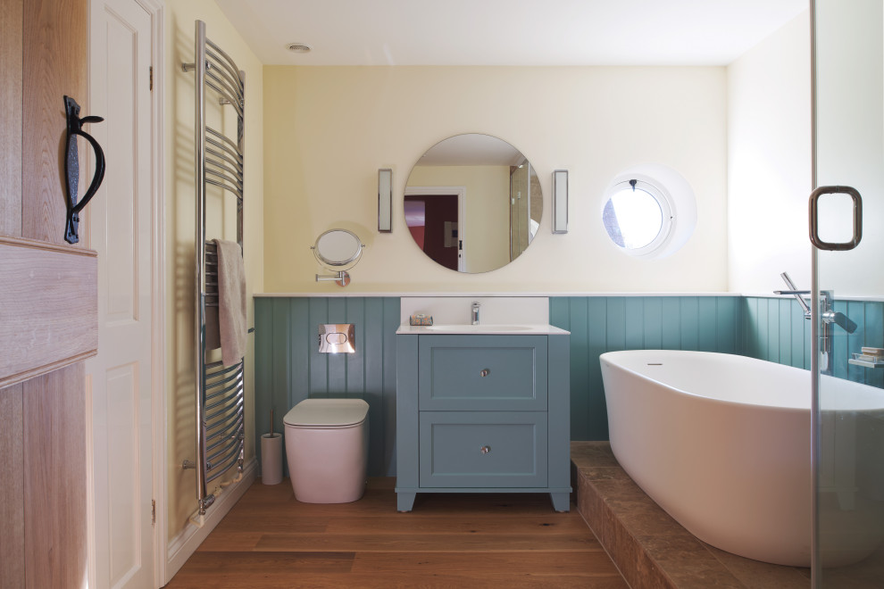 This is an example of a traditional bathroom in Wiltshire with recessed-panel cabinets, blue cabinets, a freestanding bath, a corner shower, yellow walls, medium hardwood flooring, an integrated sink, brown floors, a hinged door, white worktops, a single sink, a freestanding vanity unit and wainscoting.