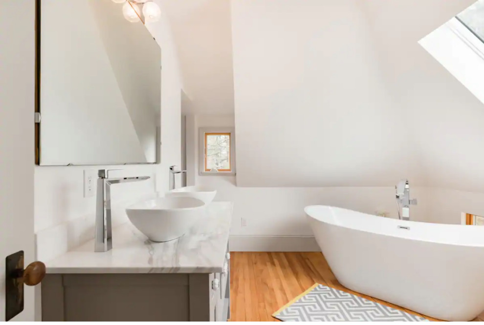 Small midcentury ensuite bathroom in Portland Maine with recessed-panel cabinets, grey cabinets, a freestanding bath, a two-piece toilet, white walls, medium hardwood flooring, a vessel sink, marble worktops, white worktops, double sinks, a freestanding vanity unit and a vaulted ceiling.