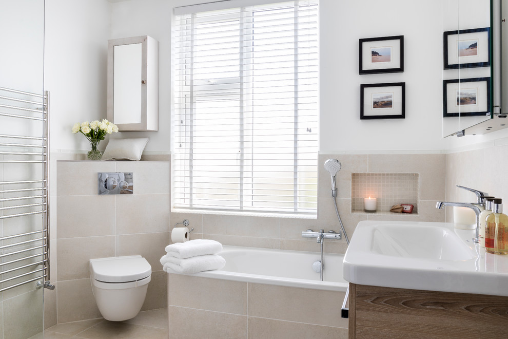 Cette photo montre une salle de bain tendance en bois clair de taille moyenne avec un placard à porte plane, une baignoire d'angle, un combiné douche/baignoire, WC suspendus, un carrelage beige, un mur blanc et un sol beige.