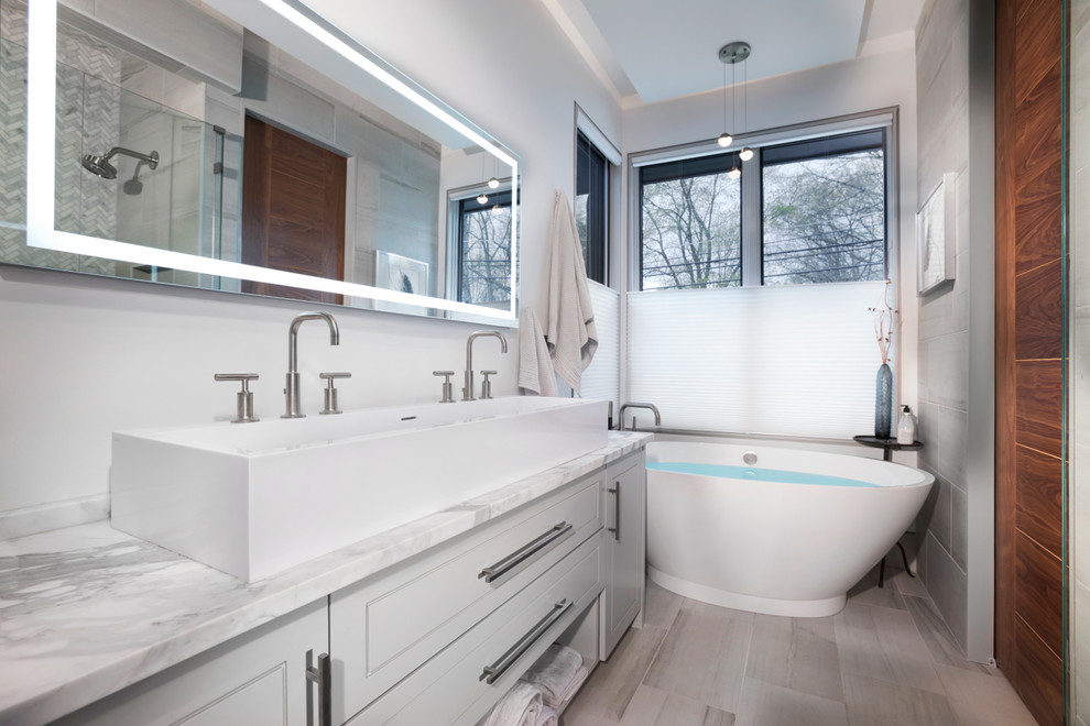 This is an example of a medium sized contemporary ensuite bathroom in Other with raised-panel cabinets, white cabinets, a freestanding bath, a corner shower, a one-piece toilet, grey tiles, marble tiles, grey walls, light hardwood flooring, a trough sink, marble worktops, beige floors, a hinged door and grey worktops.