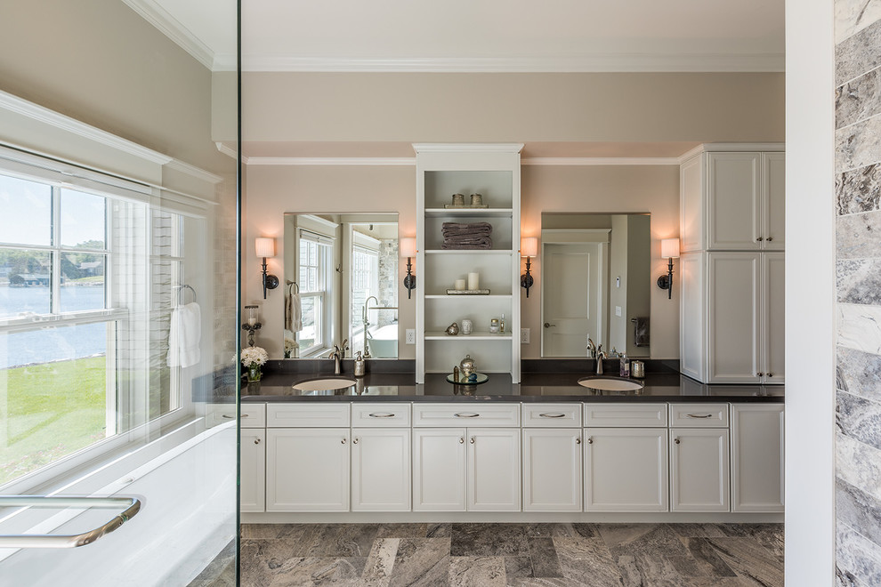 Photo of a large traditional ensuite bathroom in Minneapolis with flat-panel cabinets, white cabinets, a freestanding bath, a walk-in shower, multi-coloured tiles, stone tiles, beige walls, porcelain flooring, a submerged sink and solid surface worktops.