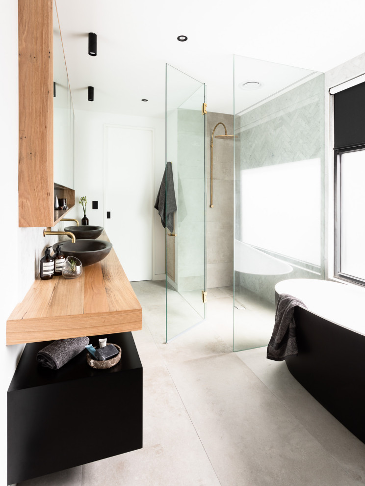 Cette photo montre une salle de bain industrielle avec des portes de placard noires, une baignoire indépendante, un carrelage gris, un mur blanc, une vasque, un plan de toilette en bois, un sol gris, un plan de toilette marron, meuble double vasque et meuble-lavabo suspendu.