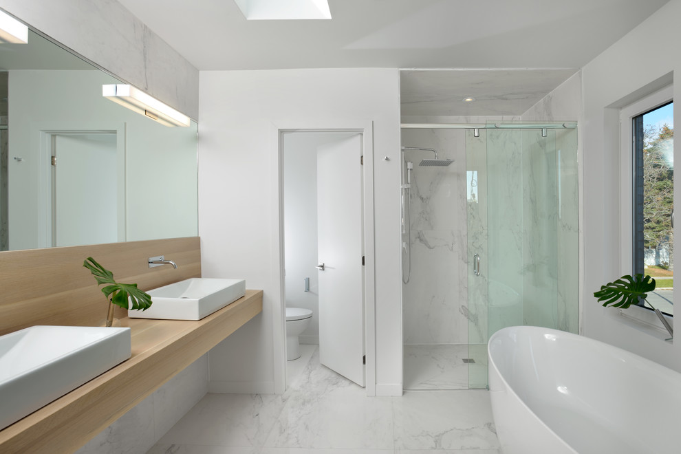 Foto de cuarto de baño contemporáneo con puertas de armario de madera clara, bañera exenta, ducha a ras de suelo, paredes blancas, suelo de mármol, lavabo sobreencimera y encimera de madera