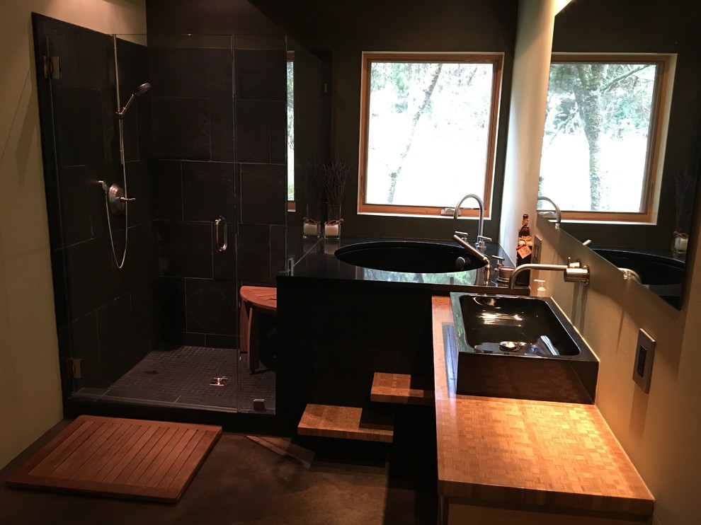 Bild på ett mellanstort industriellt badrum, med öppna hyllor, skåp i ljust trä, ett undermonterat badkar, en hörndusch, en toalettstol med separat cisternkåpa, svart kakel, stenkakel, gröna väggar, betonggolv, ett fristående handfat och granitbänkskiva