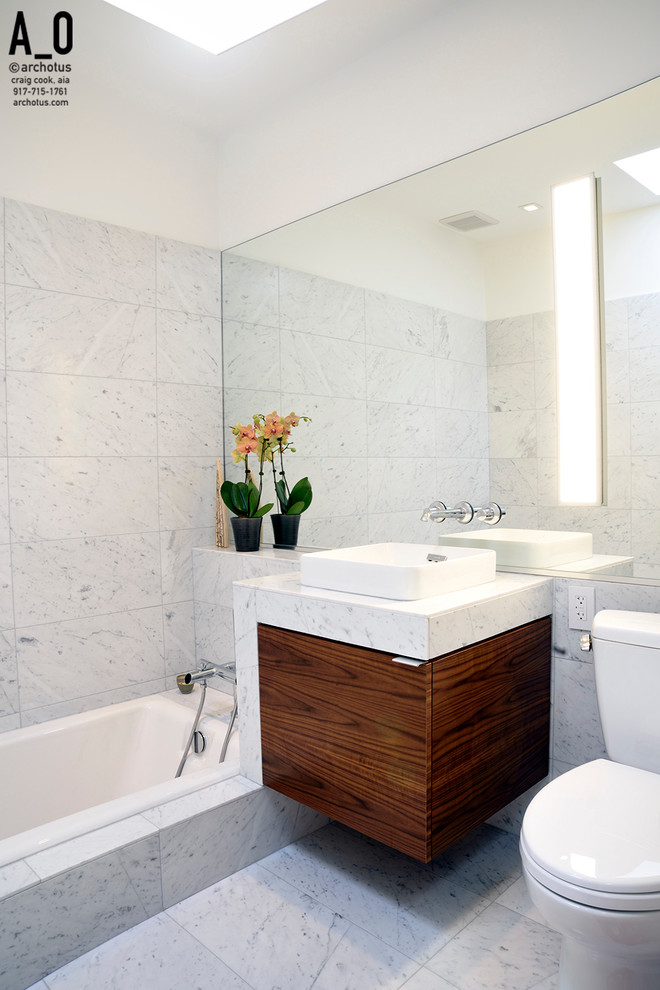 Cette photo montre une salle d'eau tendance en bois brun de taille moyenne avec un placard à porte plane, une baignoire en alcôve, WC séparés, un carrelage blanc, du carrelage en marbre, un mur blanc, un sol en marbre, une vasque, un plan de toilette en marbre et un sol blanc.