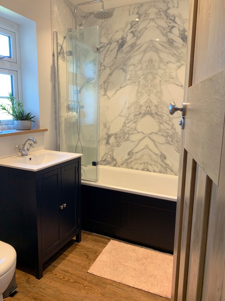 This is an example of a medium sized traditional family bathroom in Sussex with recessed-panel cabinets, blue cabinets, a built-in bath, a shower/bath combination, marble tiles, medium hardwood flooring, a console sink, brown floors, a hinged door, a single sink and a freestanding vanity unit.