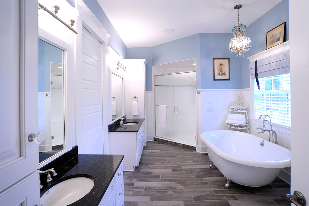 Пример оригинального дизайна: главная ванная комната: освещение в стиле неоклассика (современная классика) с ванной на ножках, серым полом, фасадами в стиле шейкер, белыми фасадами, угловым душем, синими стенами, врезной раковиной, душем с распашными дверями, черной столешницей, тумбой под две раковины, встроенной тумбой и панелями на части стены