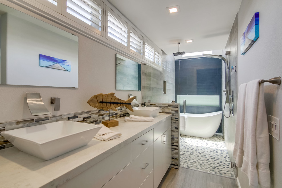 Пример оригинального дизайна: большая ванная комната в морском стиле с плоскими фасадами, белыми фасадами, отдельно стоящей ванной, душем без бортиков, синей плиткой, серой плиткой, серыми стенами, настольной раковиной, серым полом, душем с распашными дверями и белой столешницей